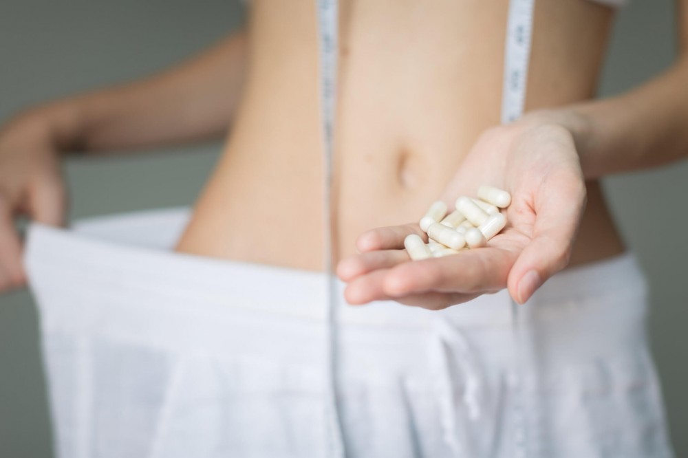 A woman showing the weight she loss with her oversize pants and holding pills.